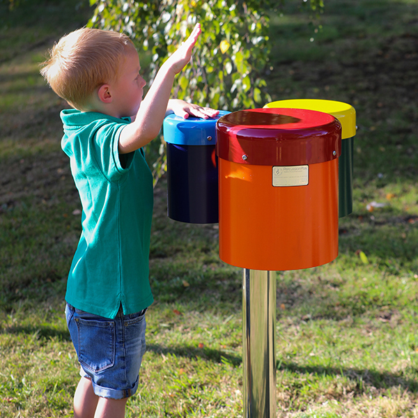 small boy reaching up and playing a colourful set of three outdoor drums mounted on a single post in a park