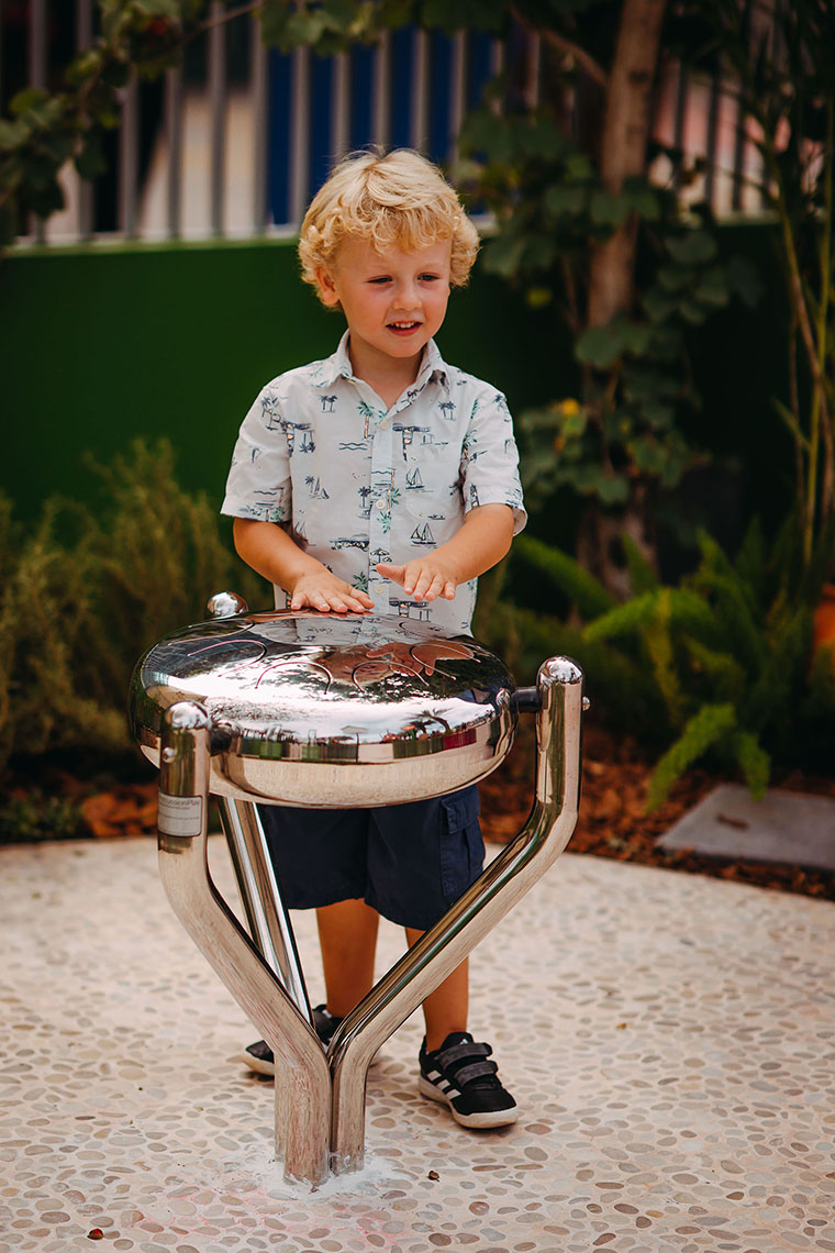 A blonde little boy playing a stainless steel tongue drum in a music park or playground