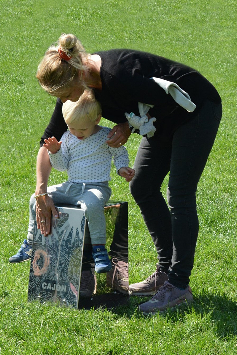 Mother standing over a child sat on a stainless steel outdoor cajon drum in a music park