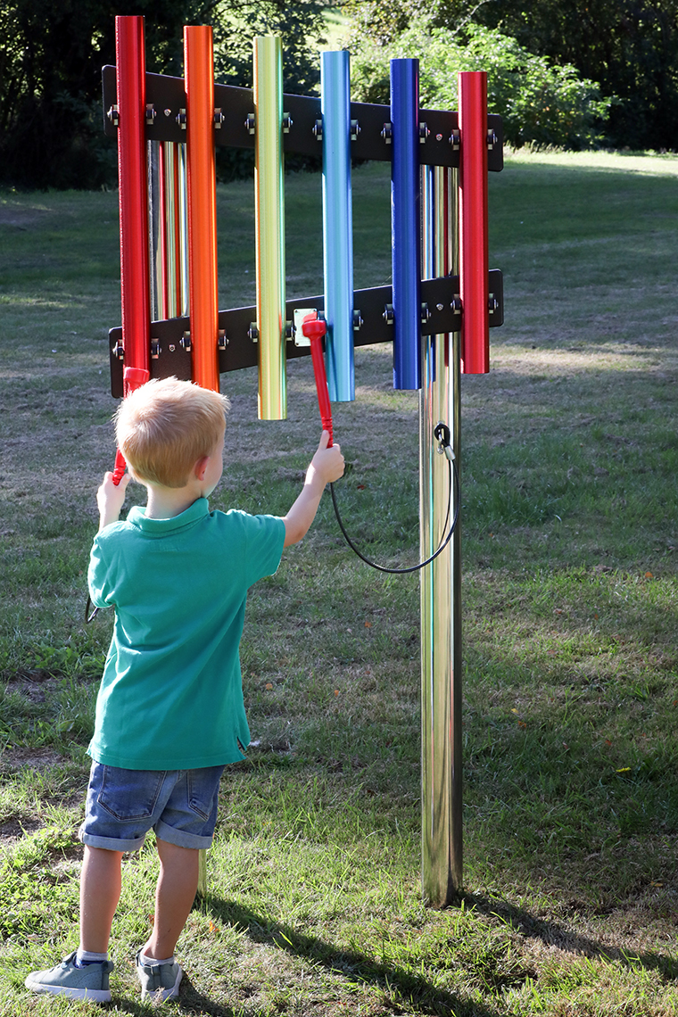 A little blonde boy playing on an outdoor musical instrument of five rainbow coloured chimes on a stainless steel post