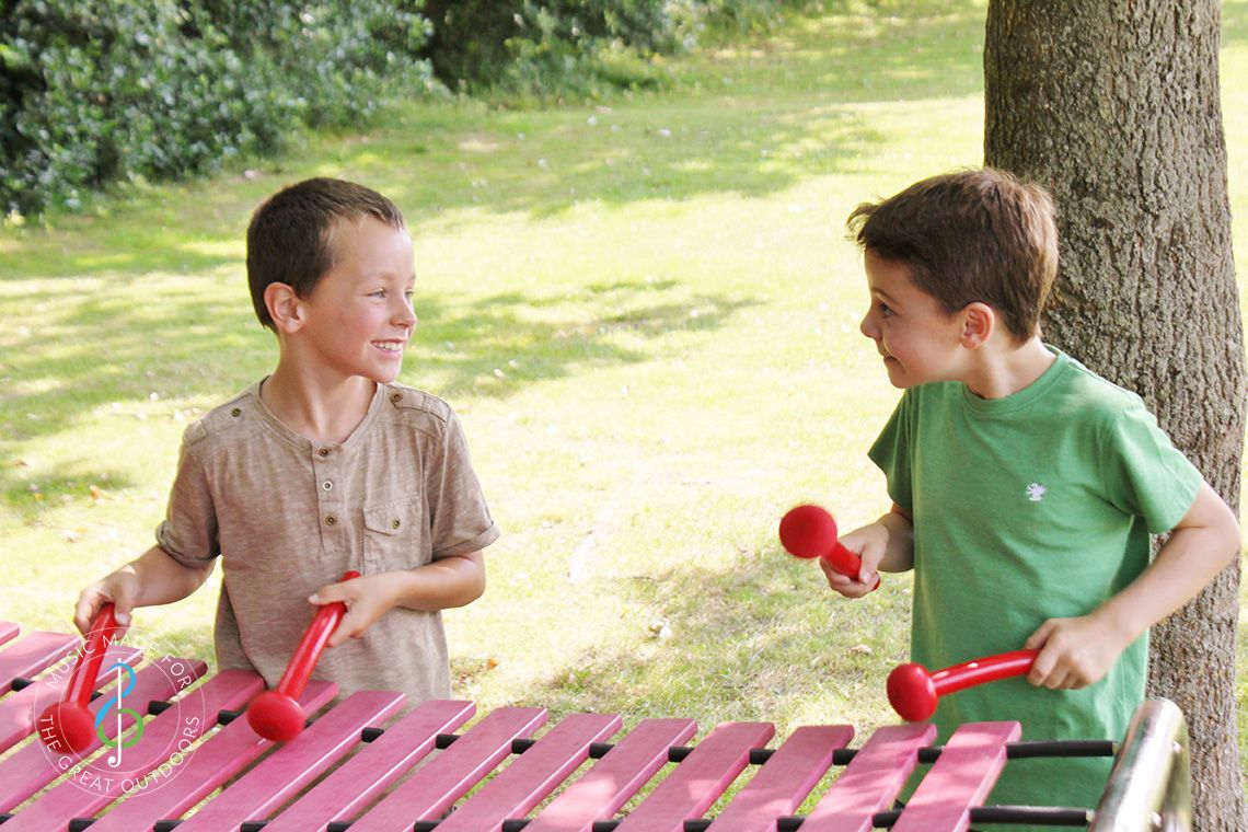 two boys laughing and playing a large outdoor marimba xylophone in a play park