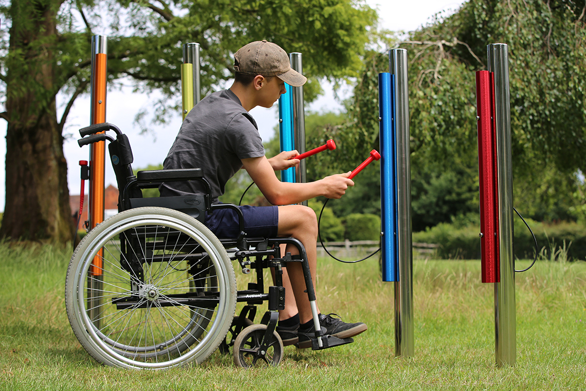 a young teenage boy in a wheelchair playing rainbow colored Calypson Chimes in a musical playground