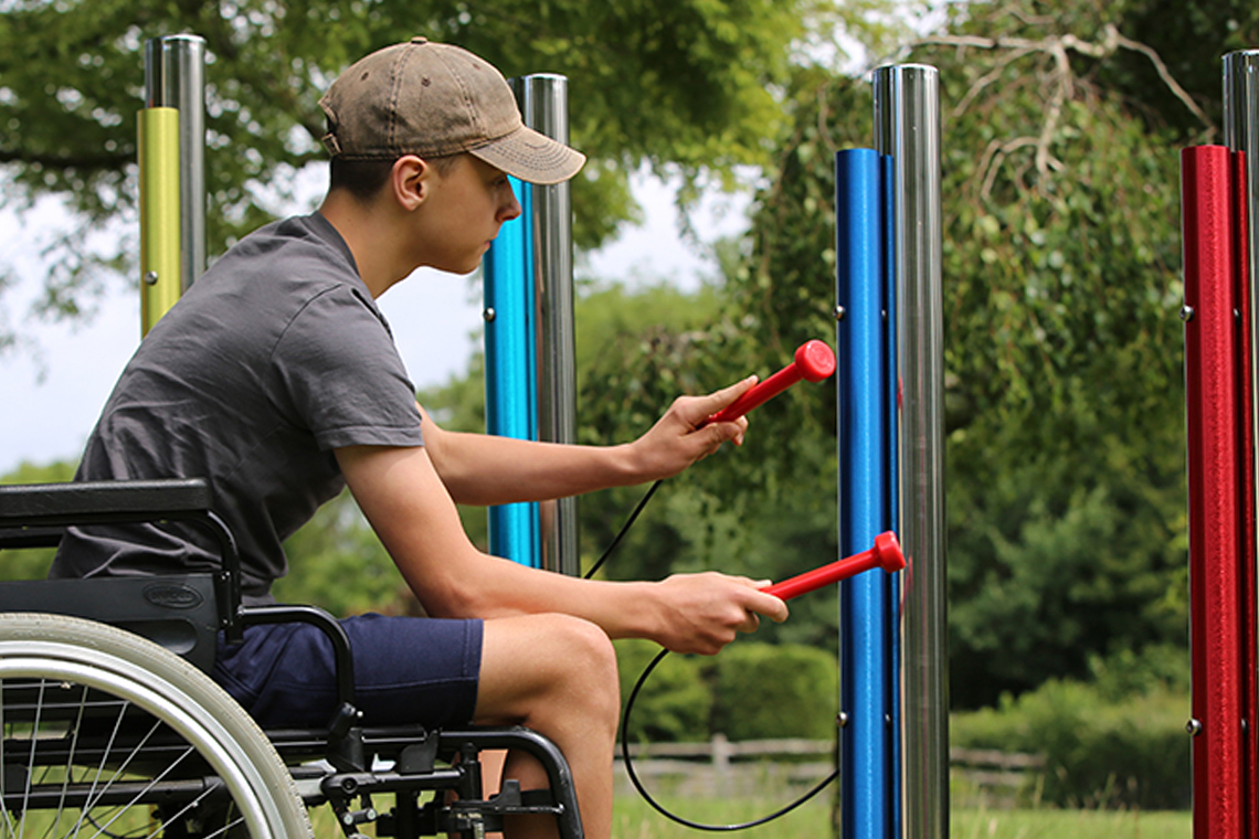 a young man in a wheelchair playing on rainbow colored outdoor musical chimes