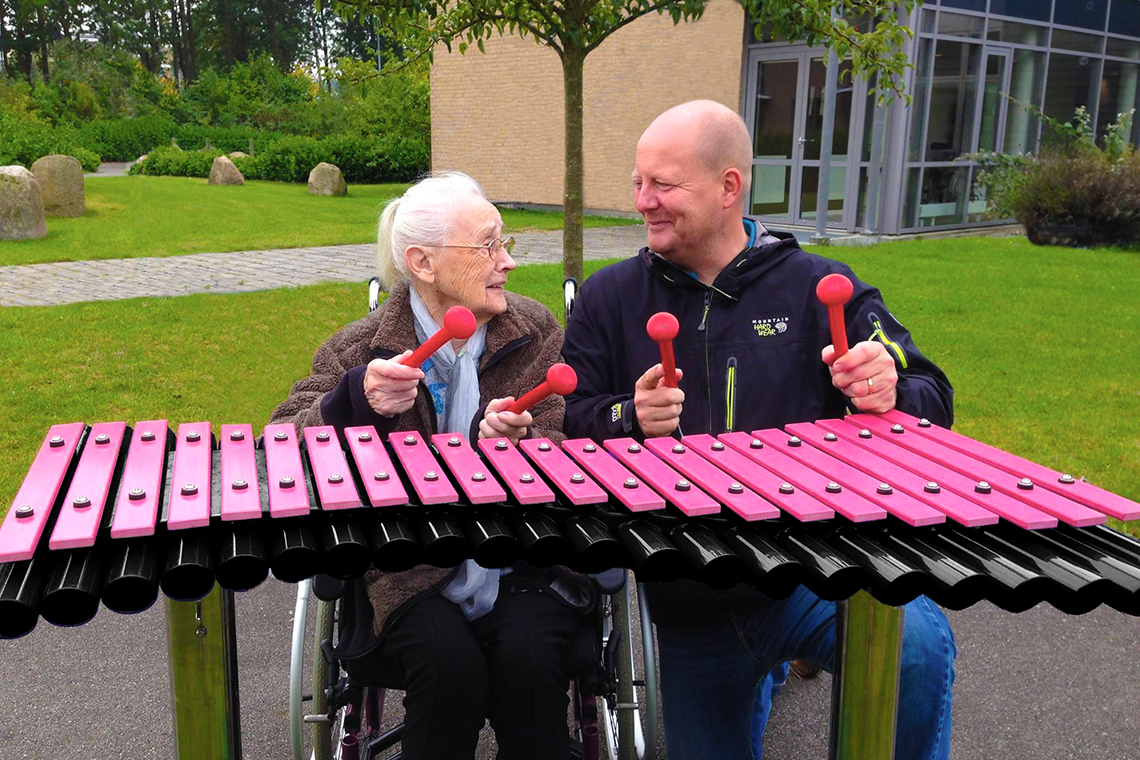 Man kneeling next to an elderly lady in a wheelchair playing an outdoor xylophone