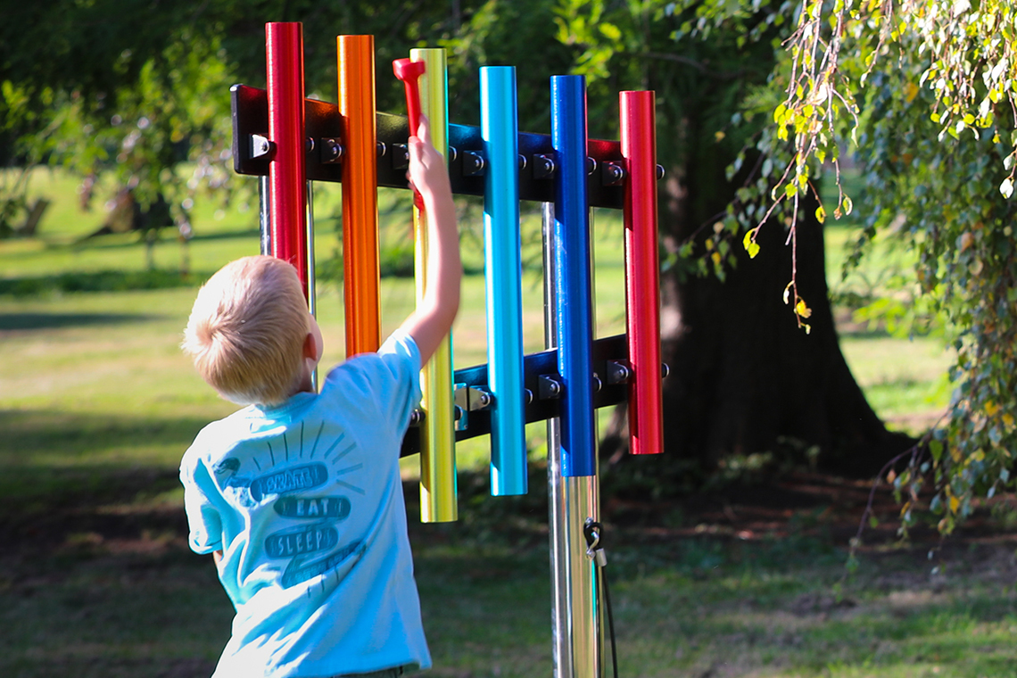 Young boy reaching up and playing a rainbow coloured set of six musical chimes in a school playground