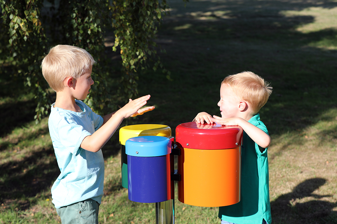 Two young brothers playing on a set of outdoor rainbow coloured drums in a playground 
