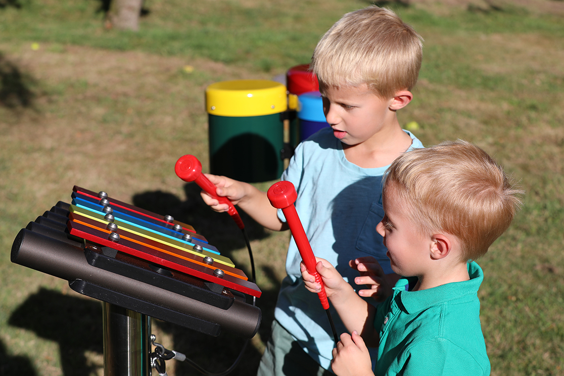 Two young brothers playing on a small rainbow coloured outdoor xylophone in a playground 