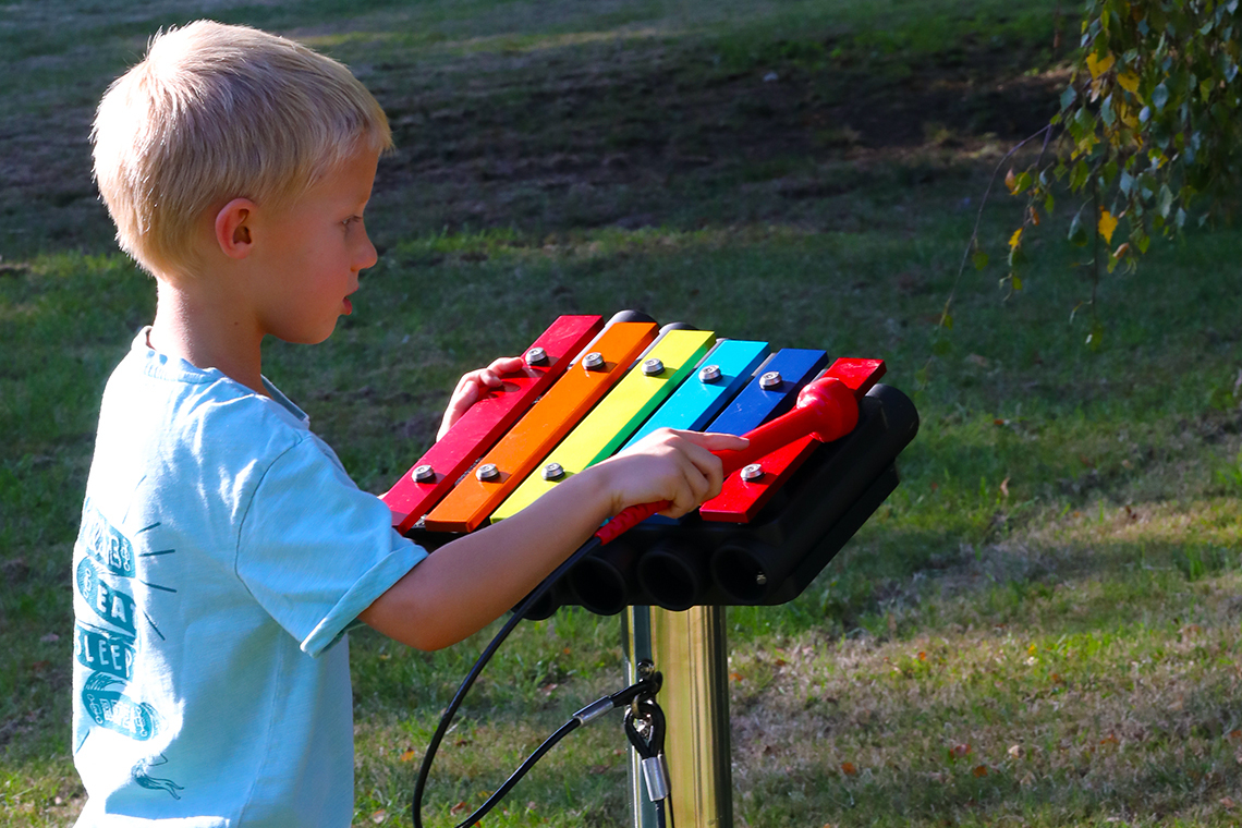 a young boy playing on a small rainbow coloured outdoor xylophone mounted on a stainless steel post in a playground