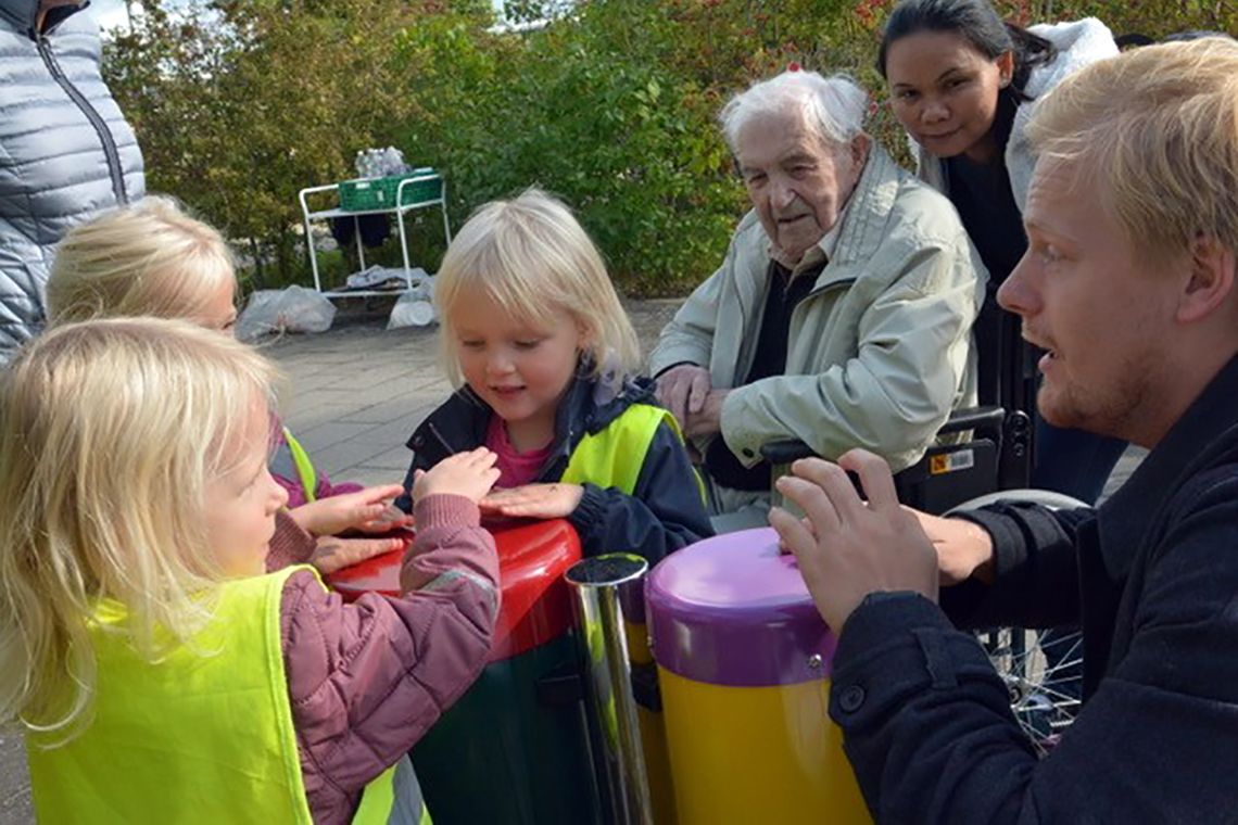 Older man in wheelchair watching three children and teacher playing outdoor conga drums in a playground