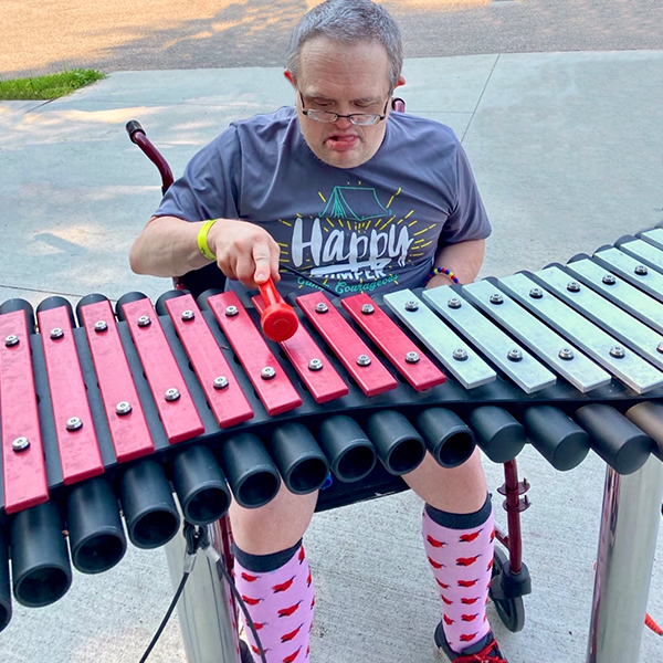 an older man with special needs and in a wheelchair playing a large outdoor xylophone in a music park at summer camp