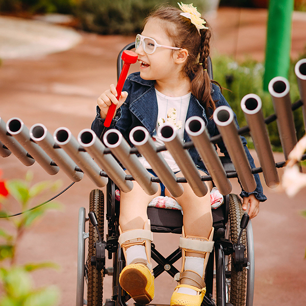 a young girl with special needs and in a wheelchair smiling and playing a large metal outdoor musical instrument in a playground