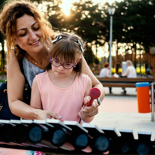 a young girl with disabilities playing a large outdoor musical instrument in the park with her mother or carer