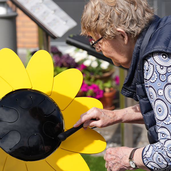 an older female adult playing an outdoor tongue drum that looks like a large sunflower