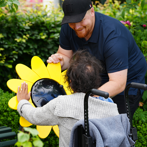A carer and a lady with disabilities and using a wheelchair is playing a large outdoor musical drum shaped like a sunflower