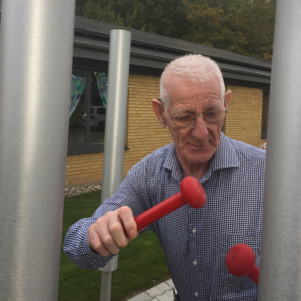 an older man hitting a tubular bell musical chime with a large red mallet