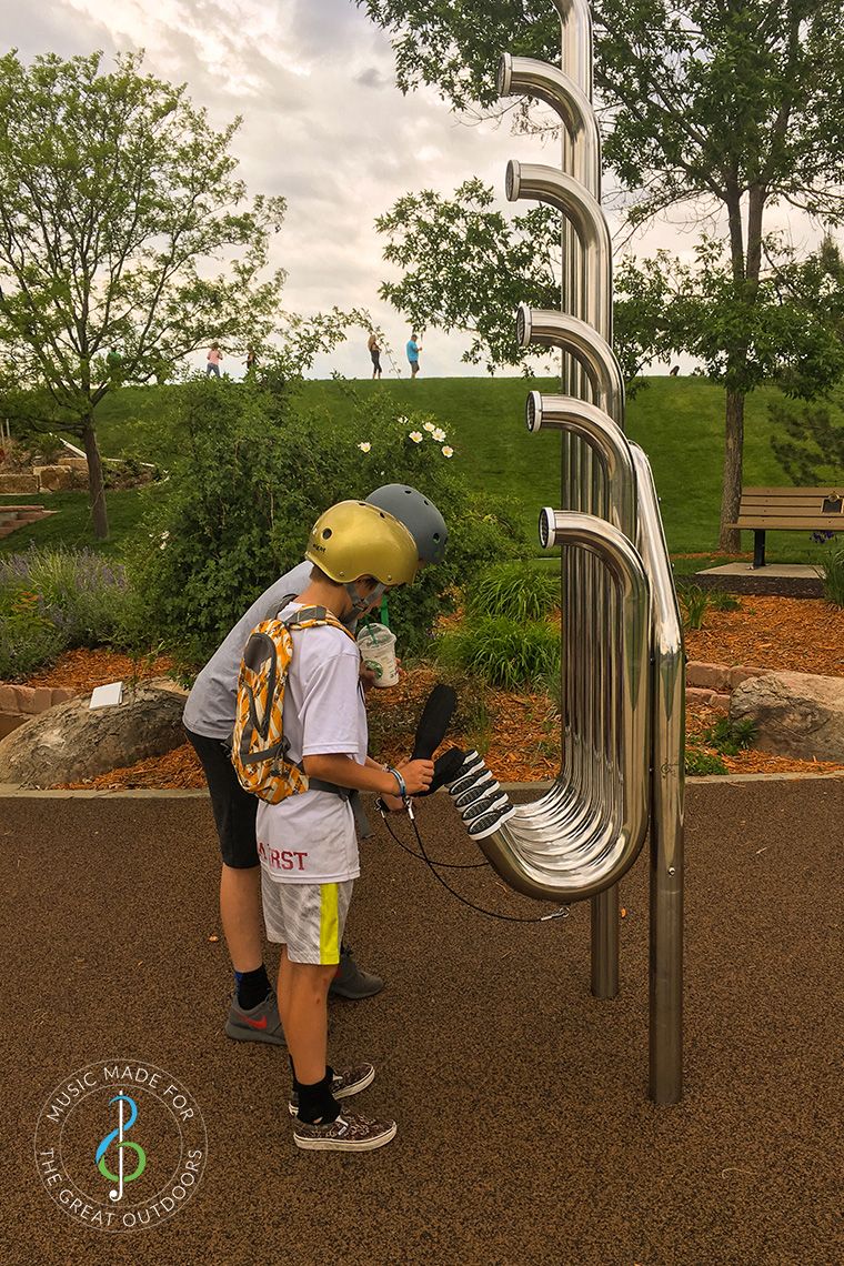 two skater boys in helmets hitting large silver outdoor aerophones or slap tubes in playground