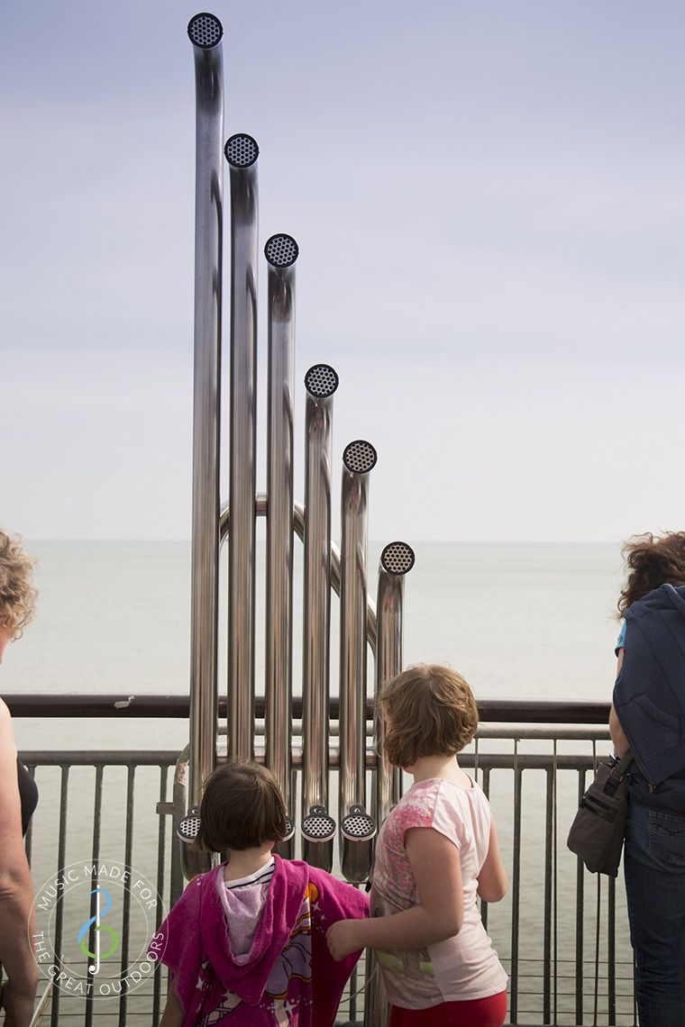 back of two girls hitting large silver outdoor aerophones or slap tubes on the end of a pier looking out to sea