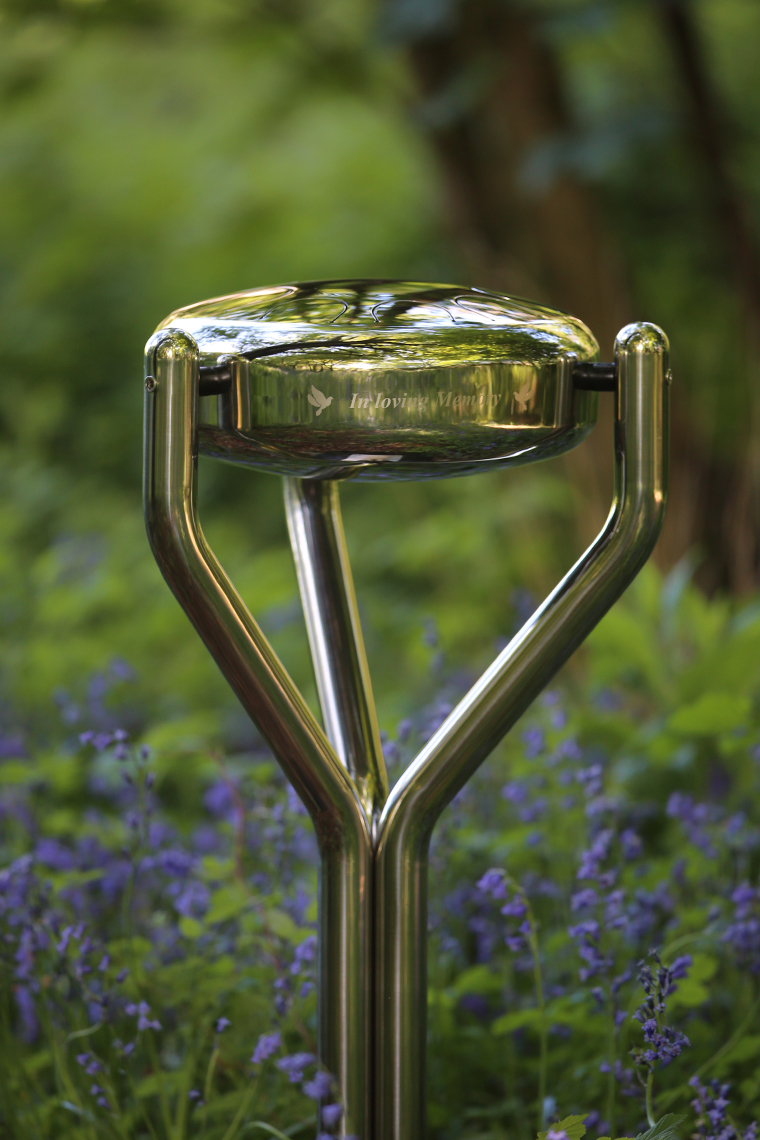 a stainless steel tongue drum engraved In Loving Memory in a memorial garden surrounded by bluebells