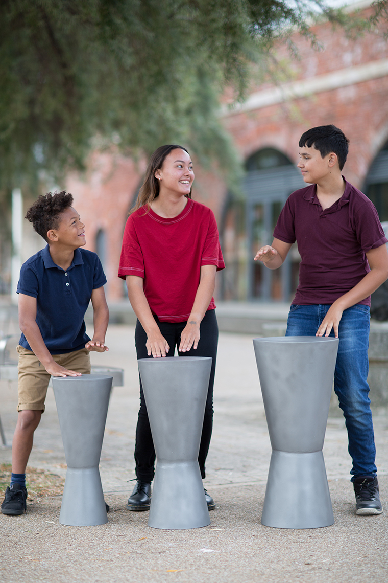 three older children playing three outdoor djembe drums with their hands in a street location