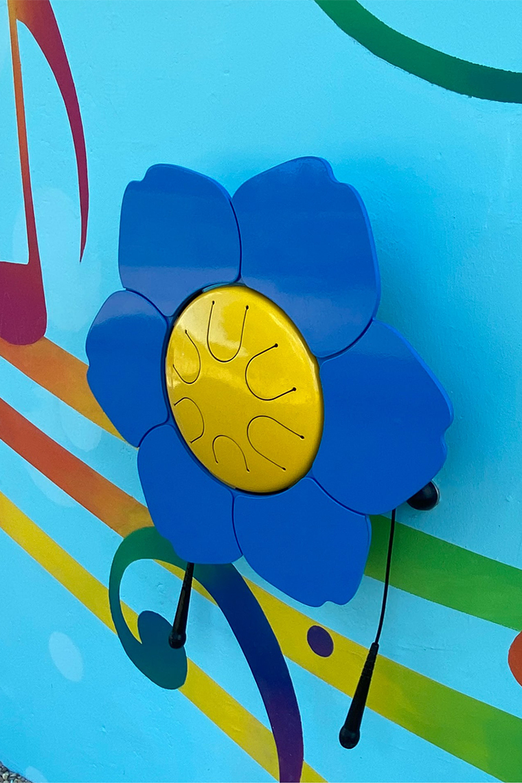 a wall mounted outdoor drum in the shape of a flower with blue petals and yellow tongue drum centre