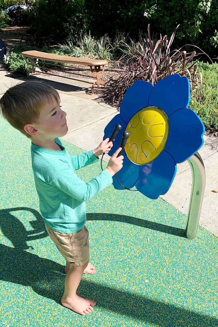 A young barefoot boy in a playground playing an outdoor drum shaped like a forget me not flower