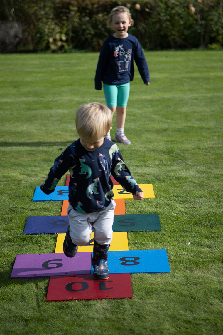a little boy jumping on an outdoor colourful musical hopscotch installed in grass with a little girl behind waiting for her turn