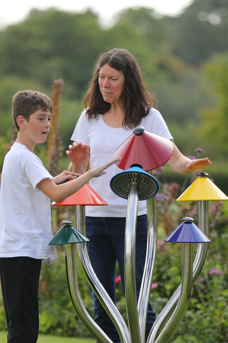 A mother and young son playing a large colorful outdoor musical instrument shaped like a cluster of mushrooms in a musical playground