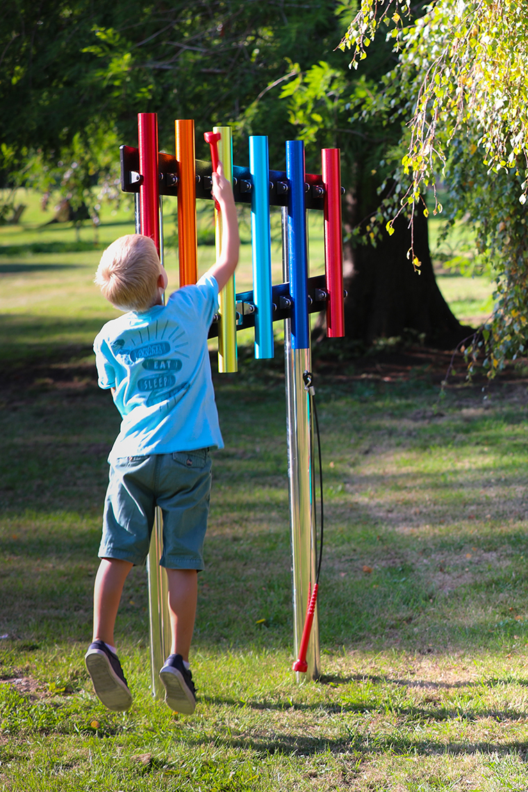 a young boy leaping up to the play the top of some rainbow coloured outdoor musical chimes in a playground 