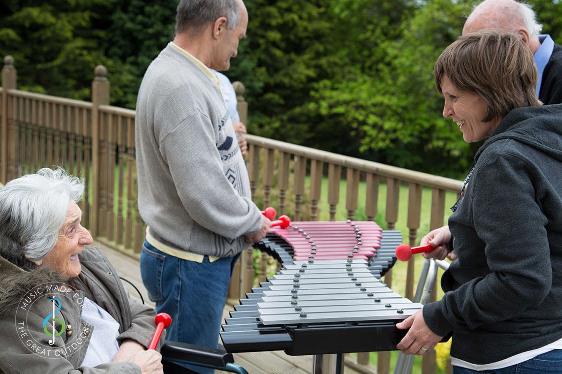 Elderly lady in a wheelchair laughing with her carer as they play on an outdoor xylophone in a care home garden