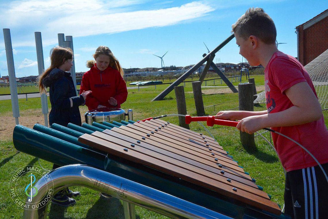 three school children playing outdoor musical instruments in school playground