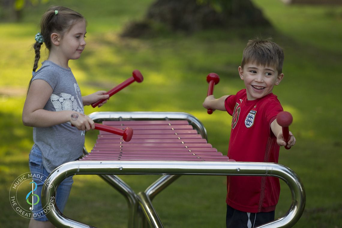 Boy and girl standing on opposite sides and playing a large outdoor marimba xylophone with red beaters