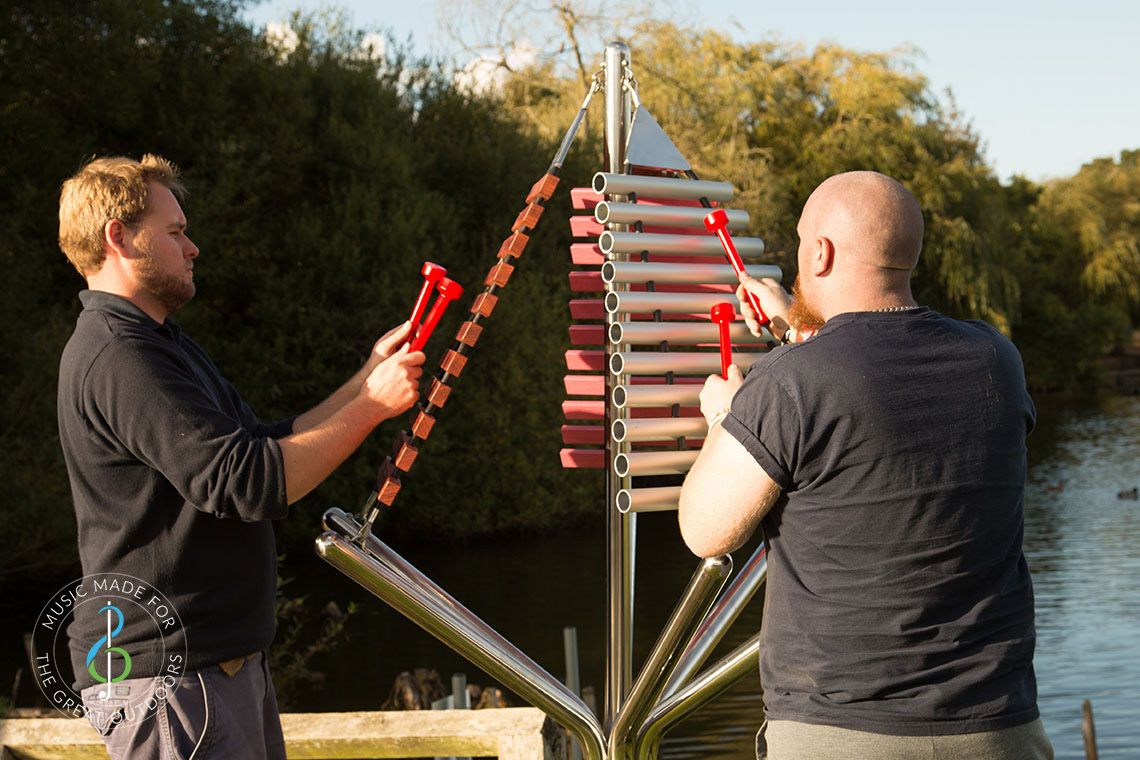 large outdoor musical instrument with one stainless steel post in ground with three vertical xylophones being played by two men
