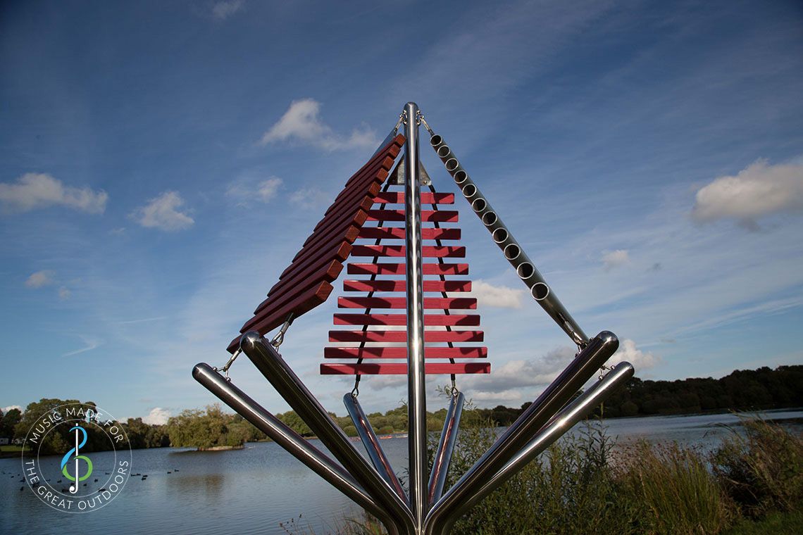 large outdoor musical instrument with one stainless steel post in ground with three vertical xylophones attached by lakeside