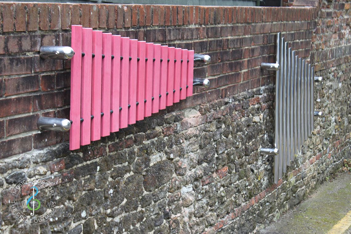one large xylophone marimba with pink notes and one set of mirrored chimes both mounted on brick wall outdoors