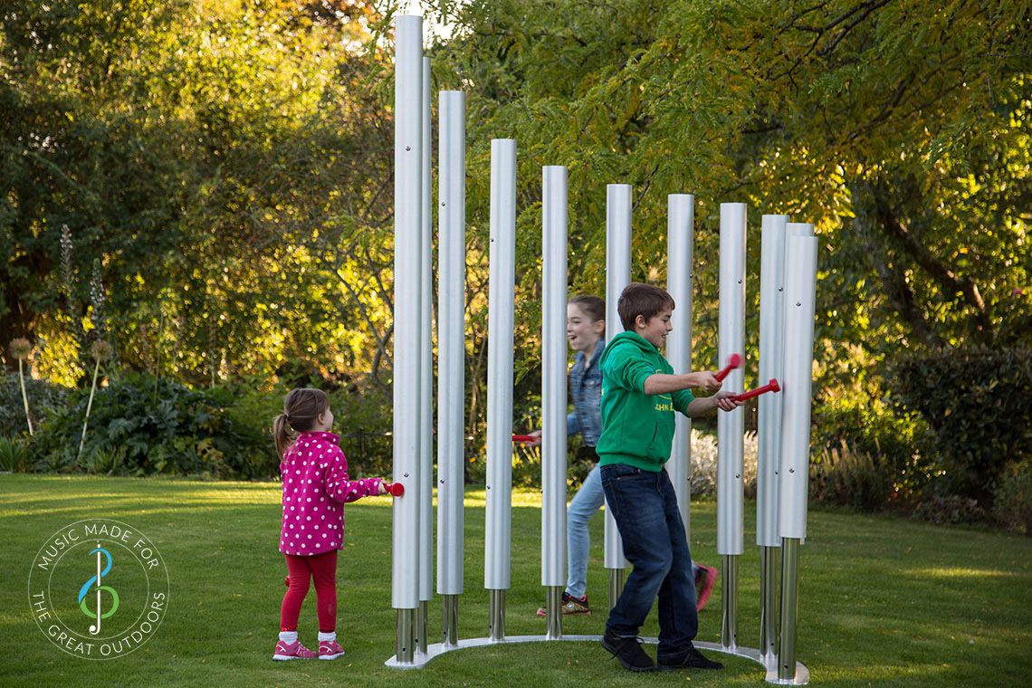 Two girls and one boy playing on 11 huge silver outdoor musical chimes set on grass