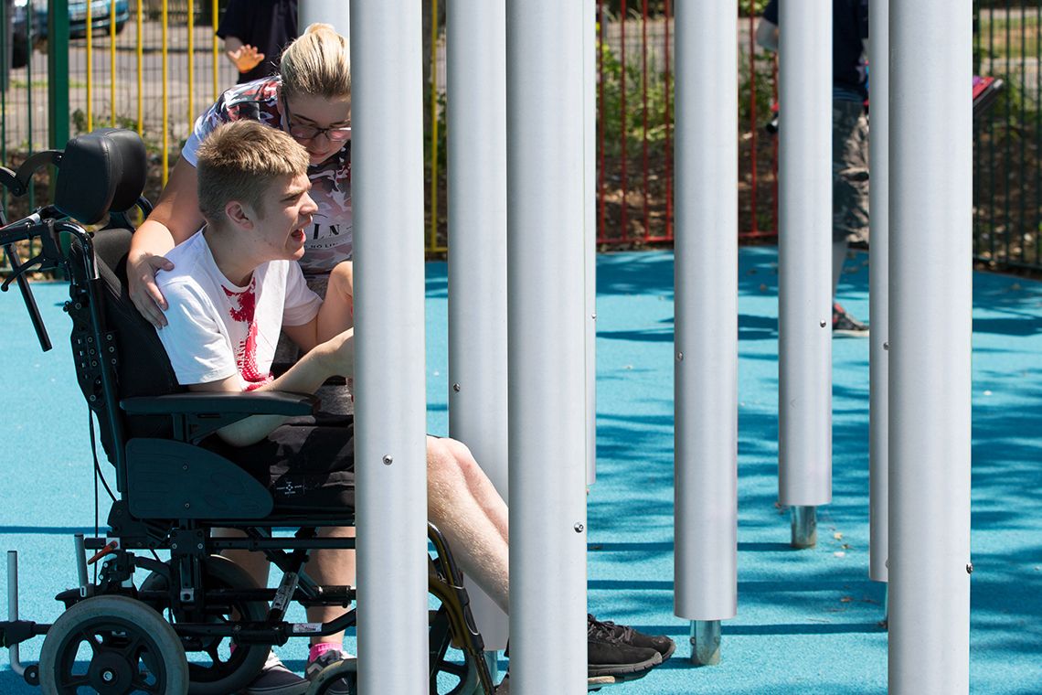 Young disabled man in a wheelchair with his carer playing on tall silver musical chimes in a school playground