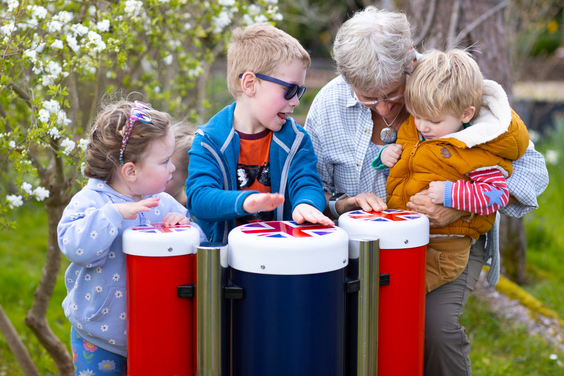 an older female adult and four young children playing outdoor conga drums in red white and blue with union flag tops