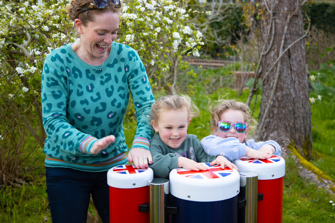 a female adult and two young children playing outdoor conga drums in red white and blue with union flag tops