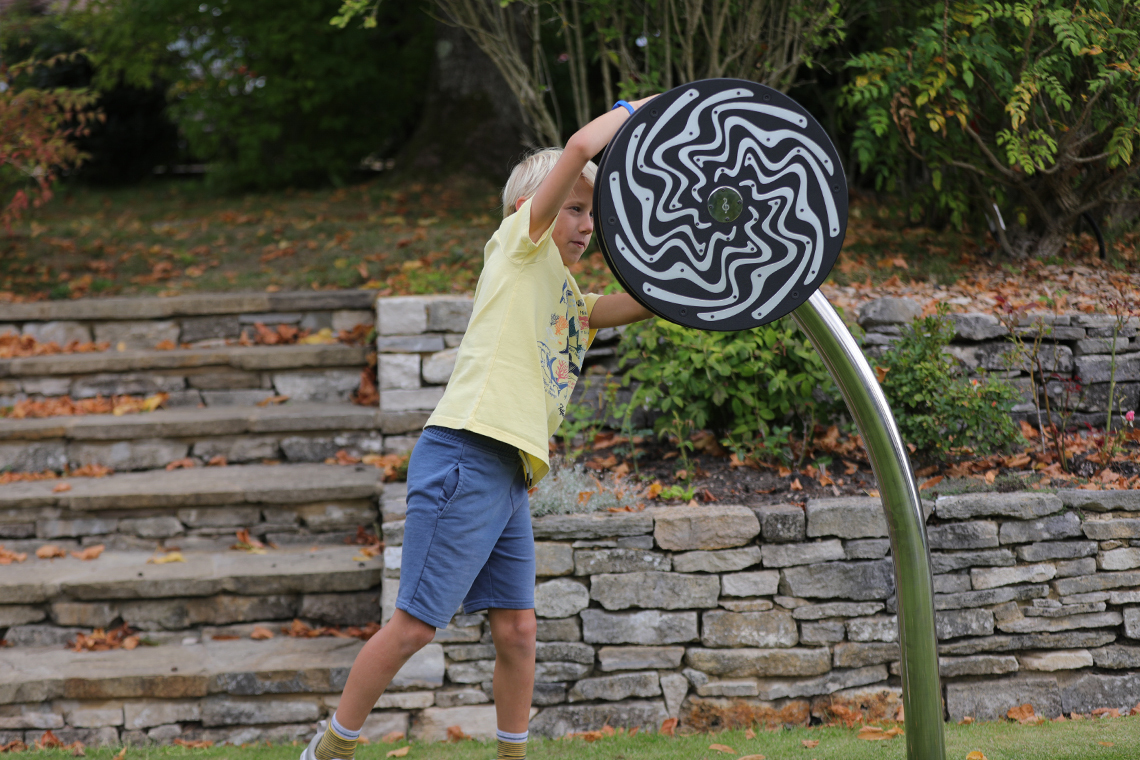 a blond boy spinning a large black and silver rain wheel in a park