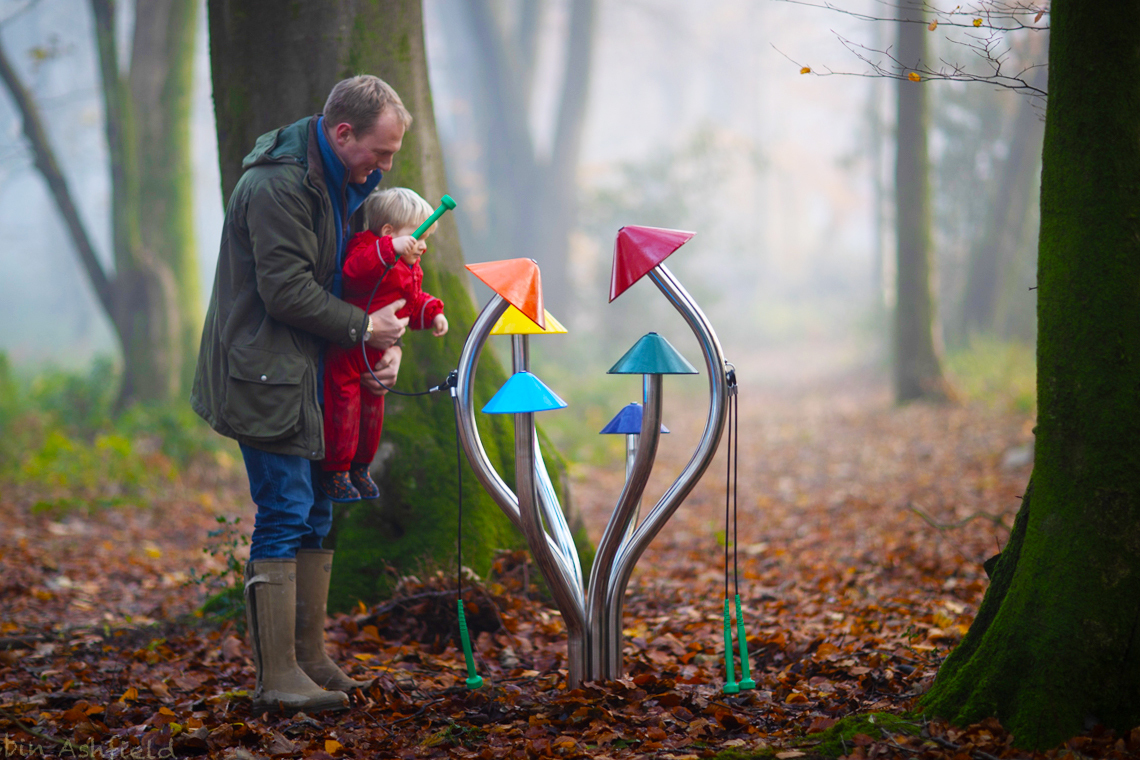 father holding his son who is wearing a red rainsuit and playing outdoor musical chimes shaped like a cluster of colorful mushrooms