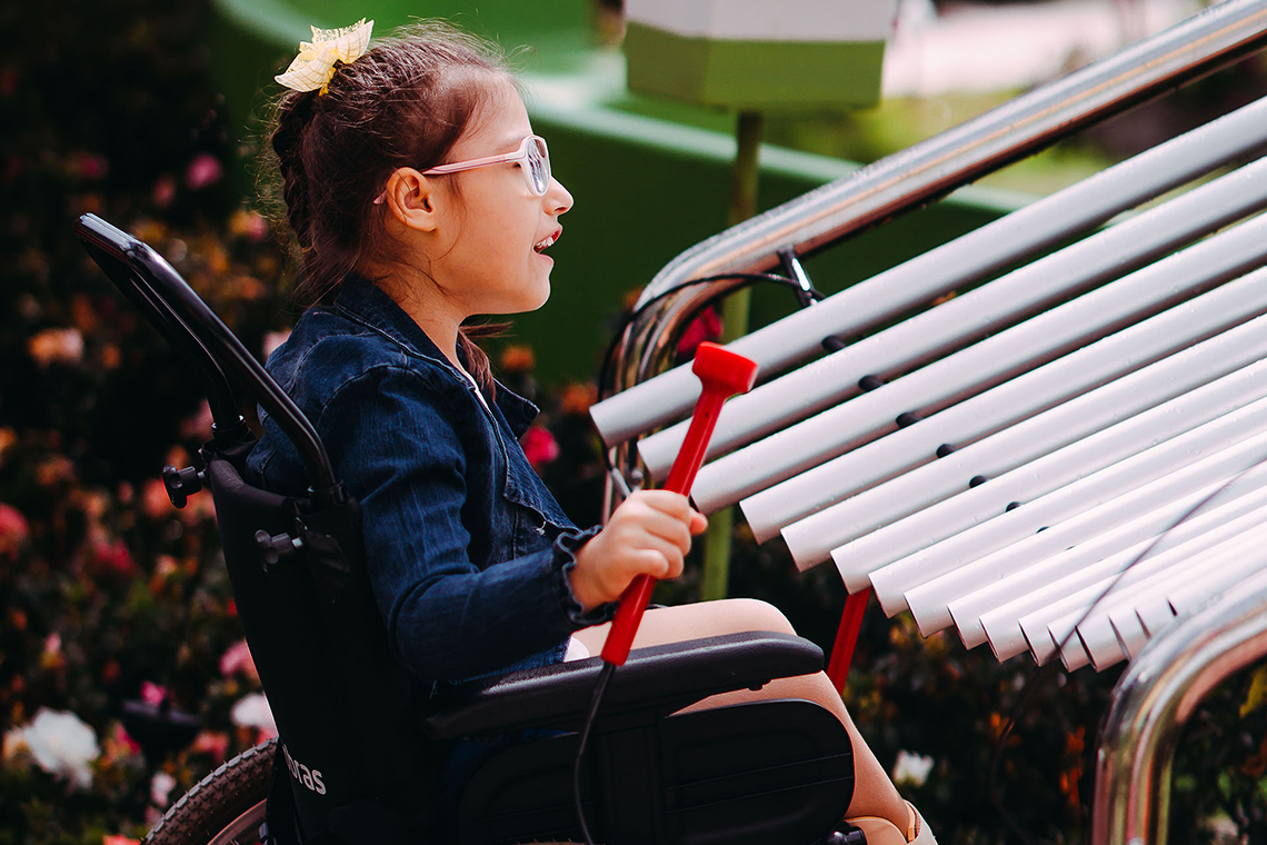 a young girl with special needs and in a wheelchair smiling and playing a large metal outdoor musical instrument in a playground