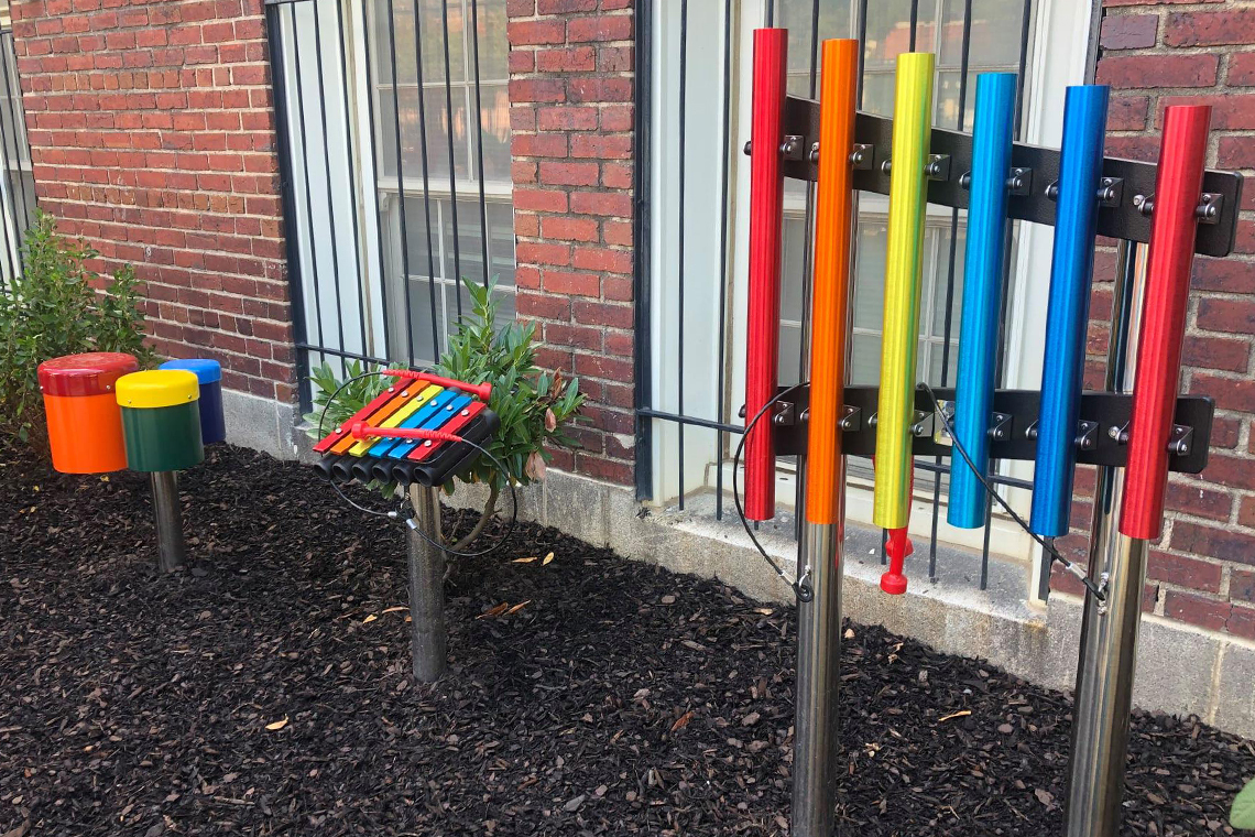 three small colorful outdoor musical instruments in a school yard; a xylophone, a set of rainbow chimes and a set of three bright bongo drums on a stainless steel post.