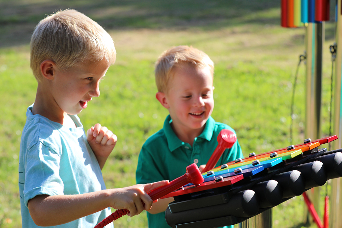Two young brothers playing on a small rainbow coloured outdoor xylophone in a playground 