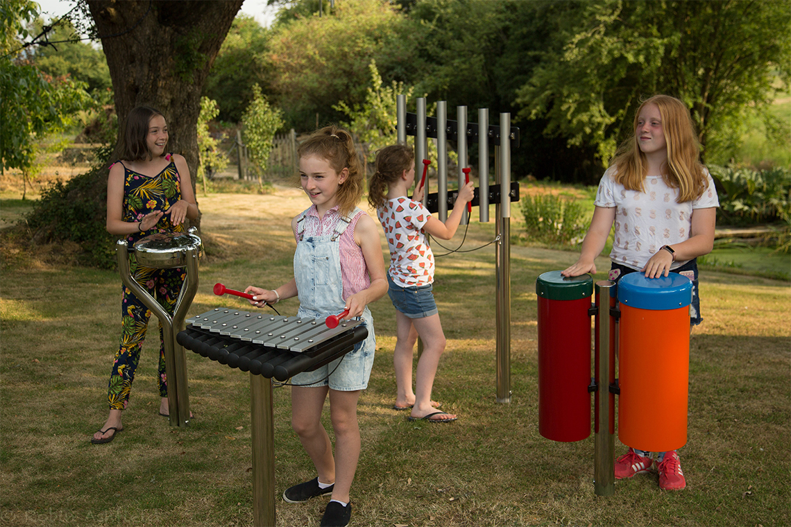 a group of four outdoor musical instruments comprising of a xylophone, chimes, pair of conga drums and stainless steel tongue drum being played by four young teenage girls