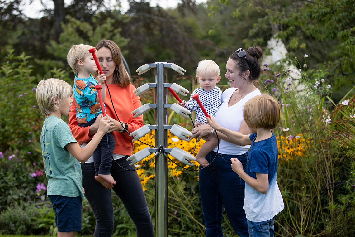 two mothers one holding a baby and with two young boys playing on an outdoor musical instrument shaped like a tree with branches made of temple blocks to be hit with red mallets