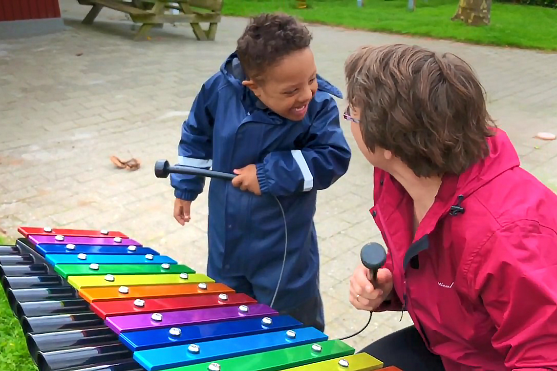 little boy with special needs playing a rainbow coloured outdoor xylophone with his teacher